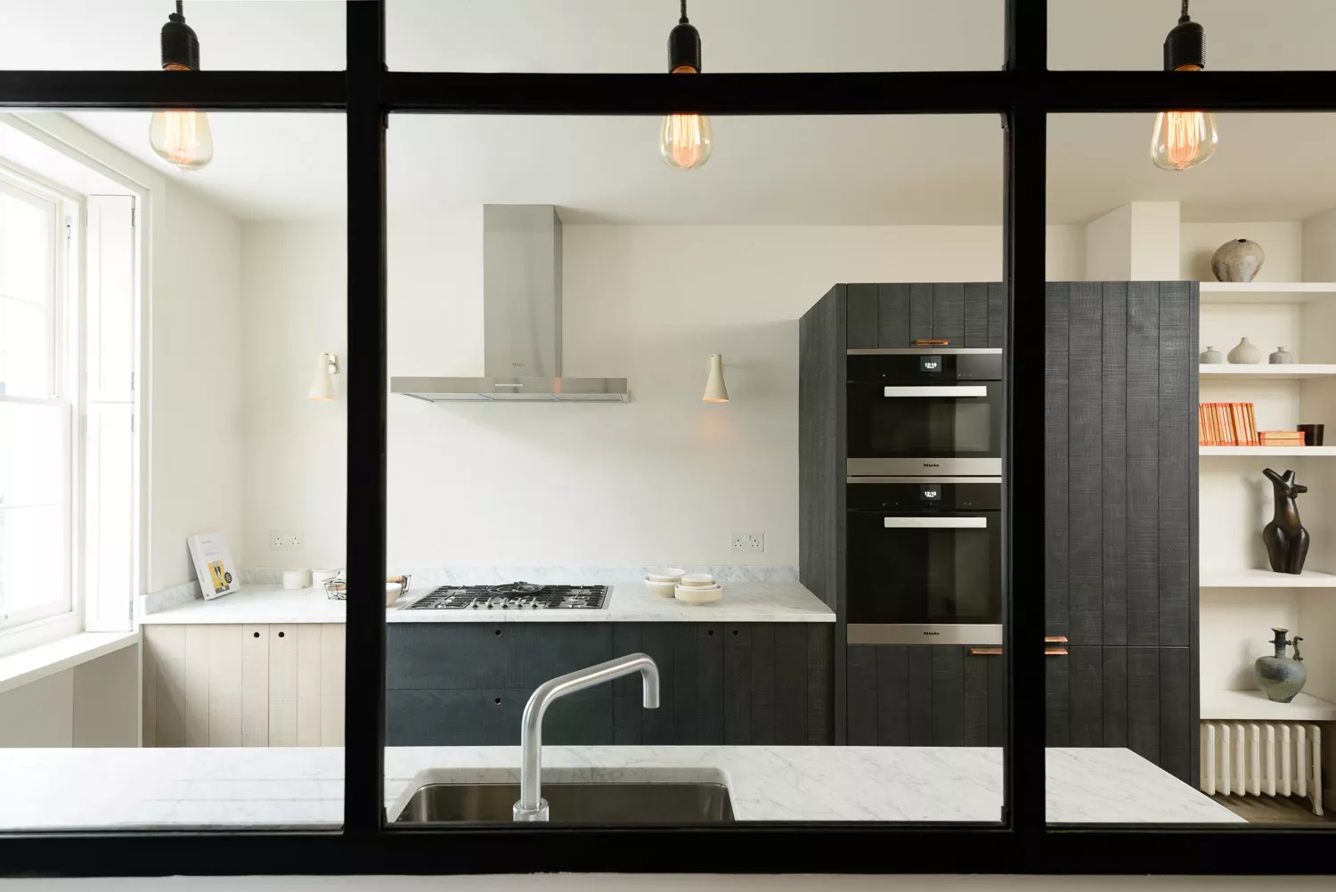 Modern minimalist London kitchen with white oak and blackened wood cabinets, industrial metal and glass partition, and stainless steel accents